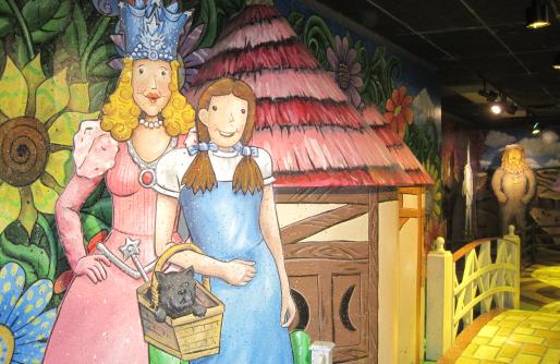 Glinda, Dorothy and Toto in front of munchkinland image of house and oversized flowers