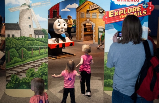 Backs of 3 young girls in pink tops entering exhibit with Thomas Engine in front of them. Side panels with train tracks, buildings and adult from back view.