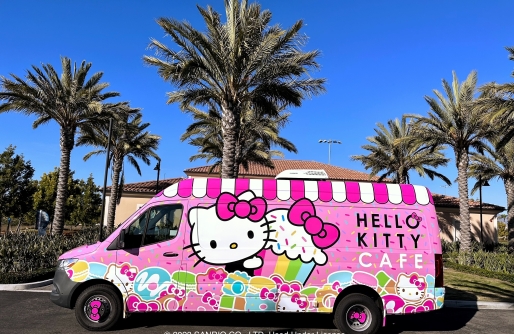 Pink Van decorated with Hello Kitty image, cupcake and other treats, parked on pavement in front of building wiht palm trees