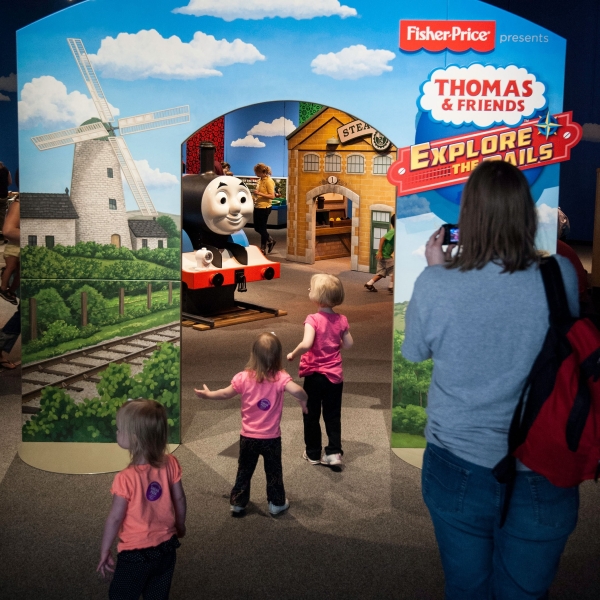 Backs of 3 young girls in pink tops entering exhibit with Thomas Engine in front of them. Side panels with train tracks, buildings and adult from back view.