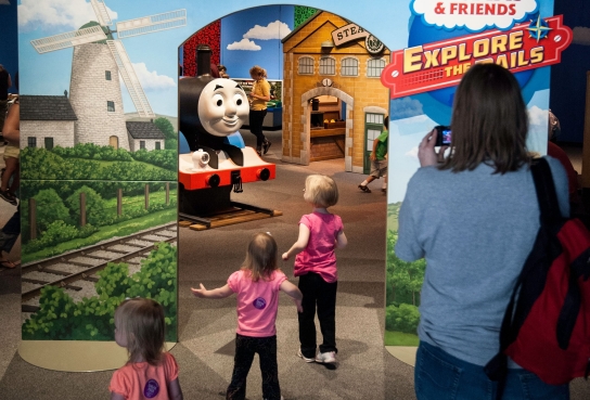 Backs of 3 young girls in pink tops entering exhibit with Thomas Engine in front of them. Side panels with train tracks, buildings and adult from back view.