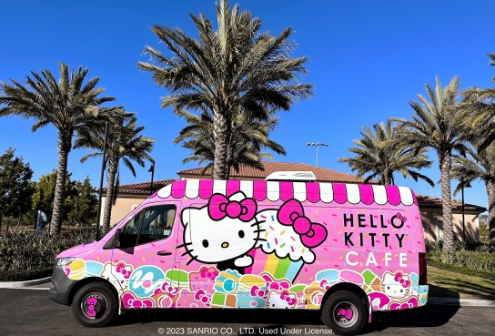 Pink Van decorated with Hello Kitty image, cupcake and other treats, parked on pavement in front of building wiht palm trees